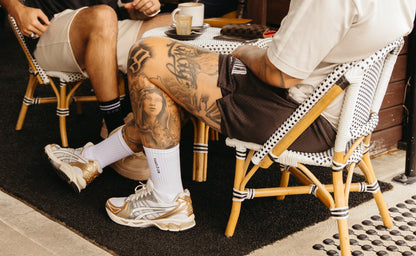 Two people sitting side by side with their legs crossed, both wearing white crew socks and sneakers. The person on the left has tattoos on their legs.