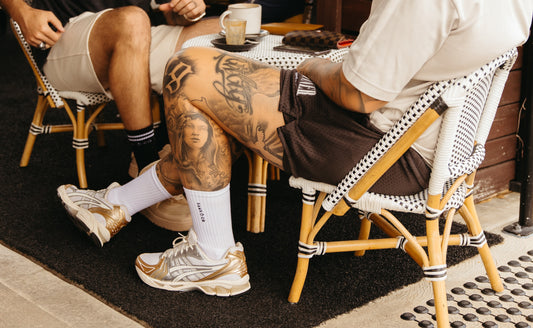 Two people sitting side by side with their legs crossed, both wearing white crew socks and sneakers. The person on the left has tattoos on their legs.