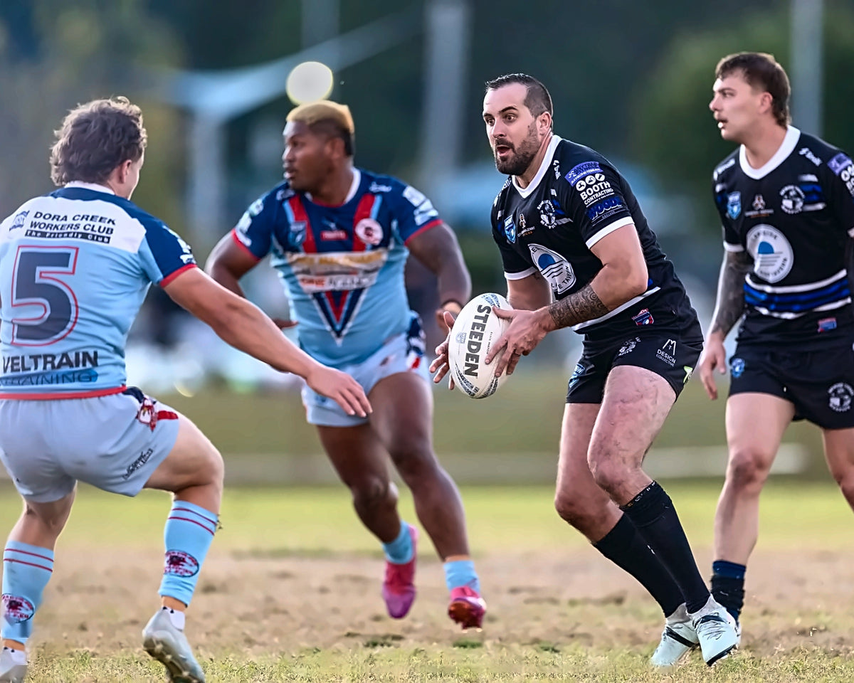 A rugby match scene with four players on a grass field. The central player in a black jersey holds the rugby ball, while teammates and opponents in blue and navy jerseys surround him.